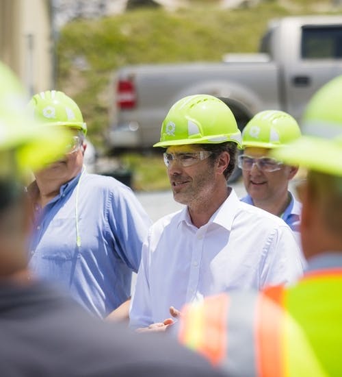 Group of men in fluorescent yellow hard hats and protective glasses, most of the group blurred with focus on man in white button-down shirt who is talking to group