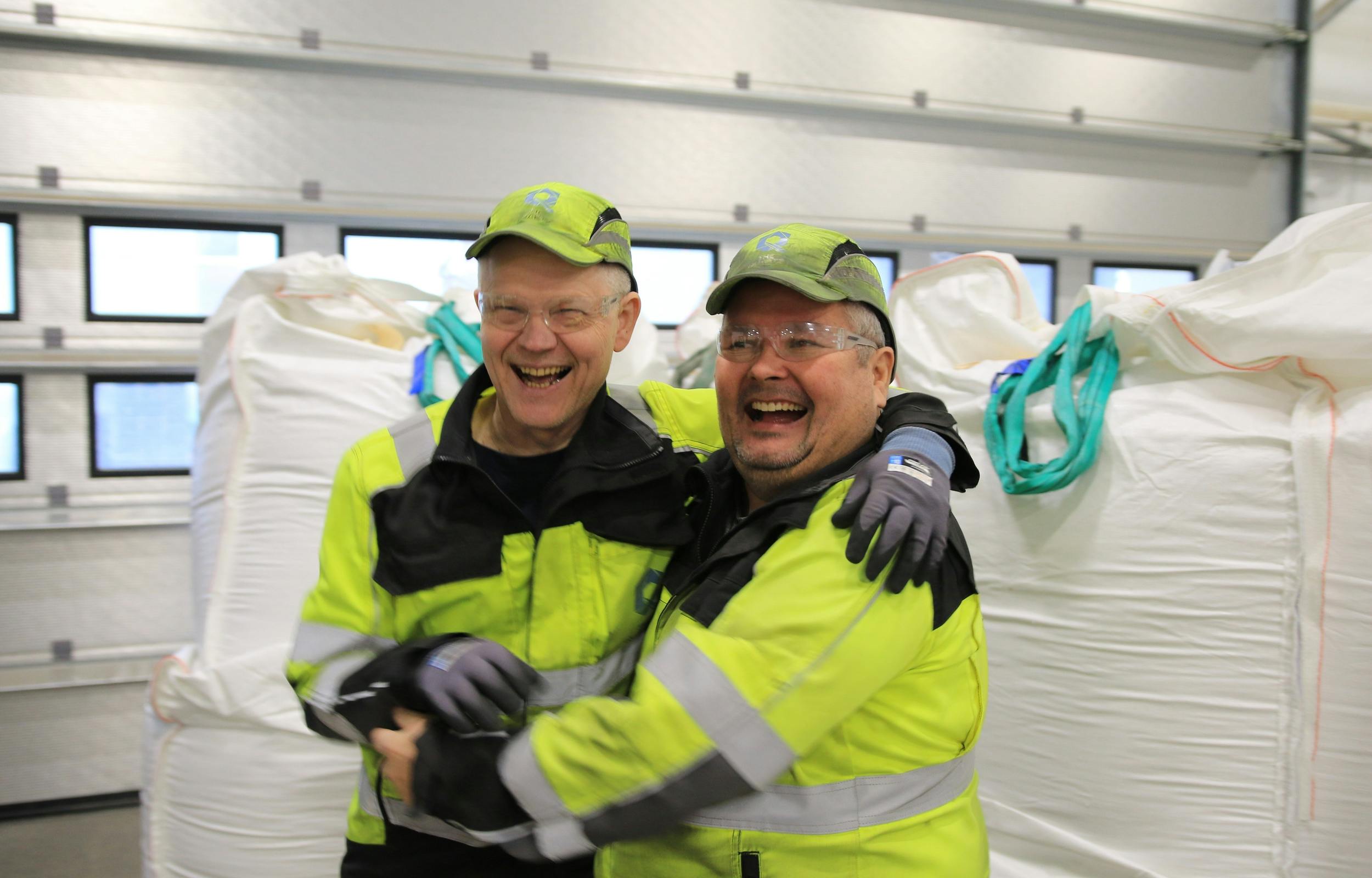 Two men who are plant workers in bright yellow safety gear hugging each other and smiling